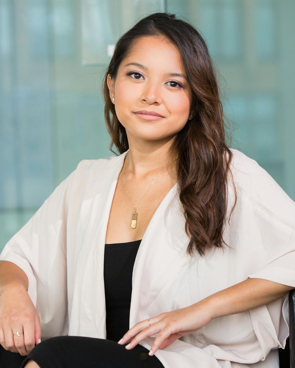 Woman in a white cardigan sits, smiling, in a bright setting; wearing a black top and gold necklace.
