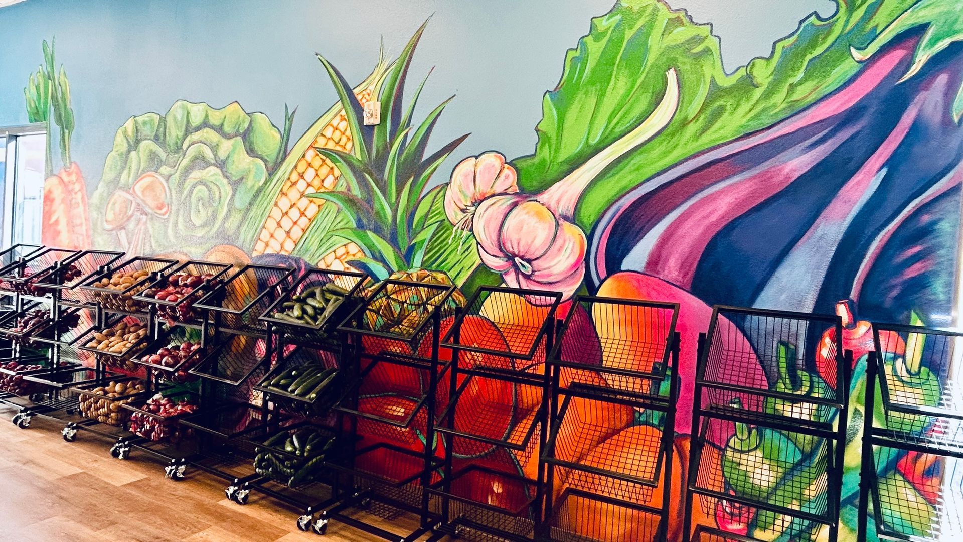 Produce bins along a wall with a vibrant mural of vegetables; carrots, lettuce, pineapple, and garlic.
