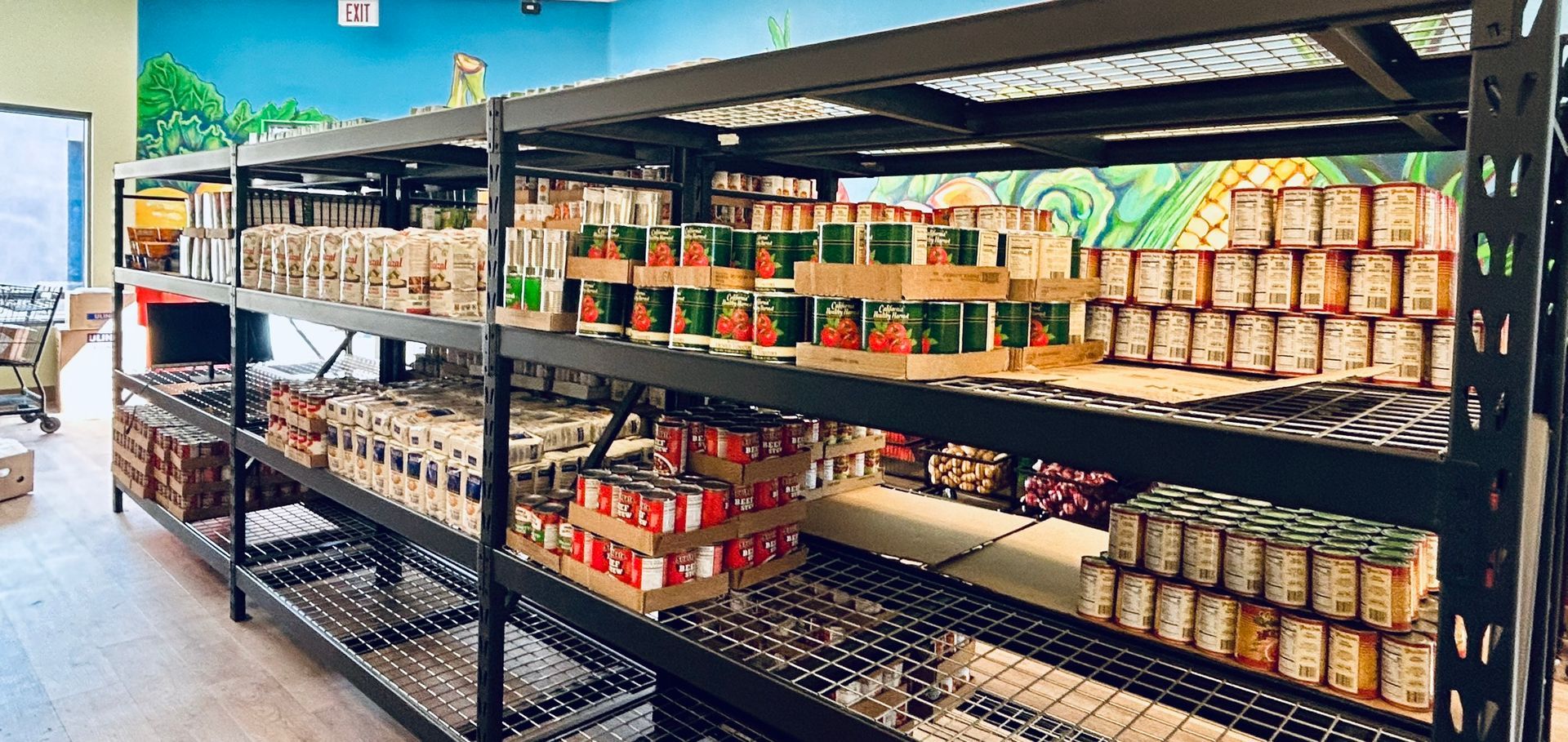 Shelves stocked with canned goods inside a food pantry with a painted mural.