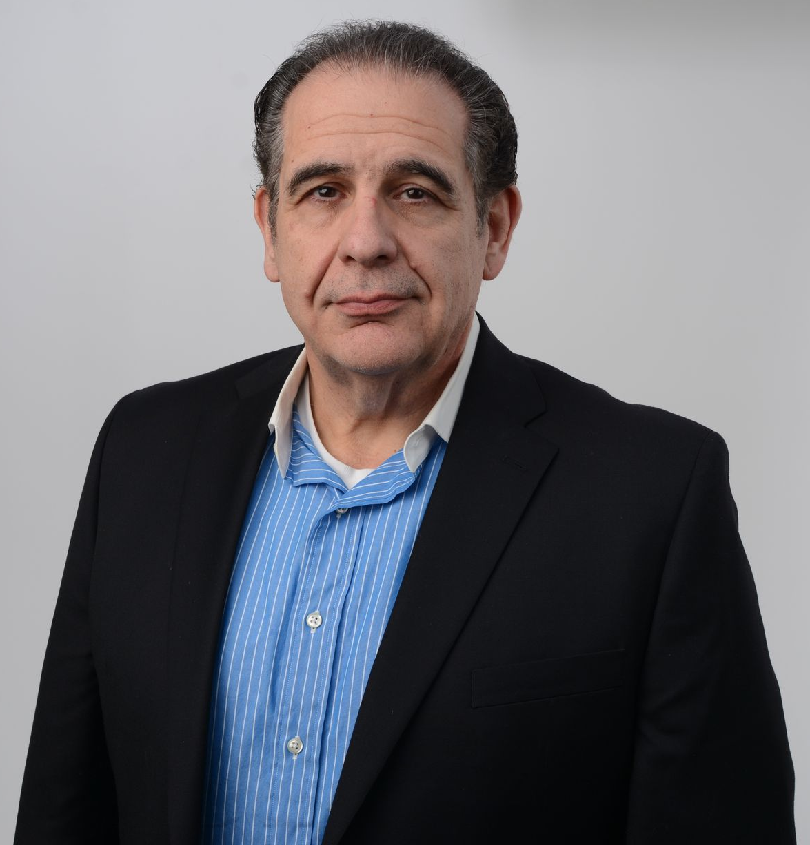 A man in a blue shirt under a black blazer looking at the camera. White background.