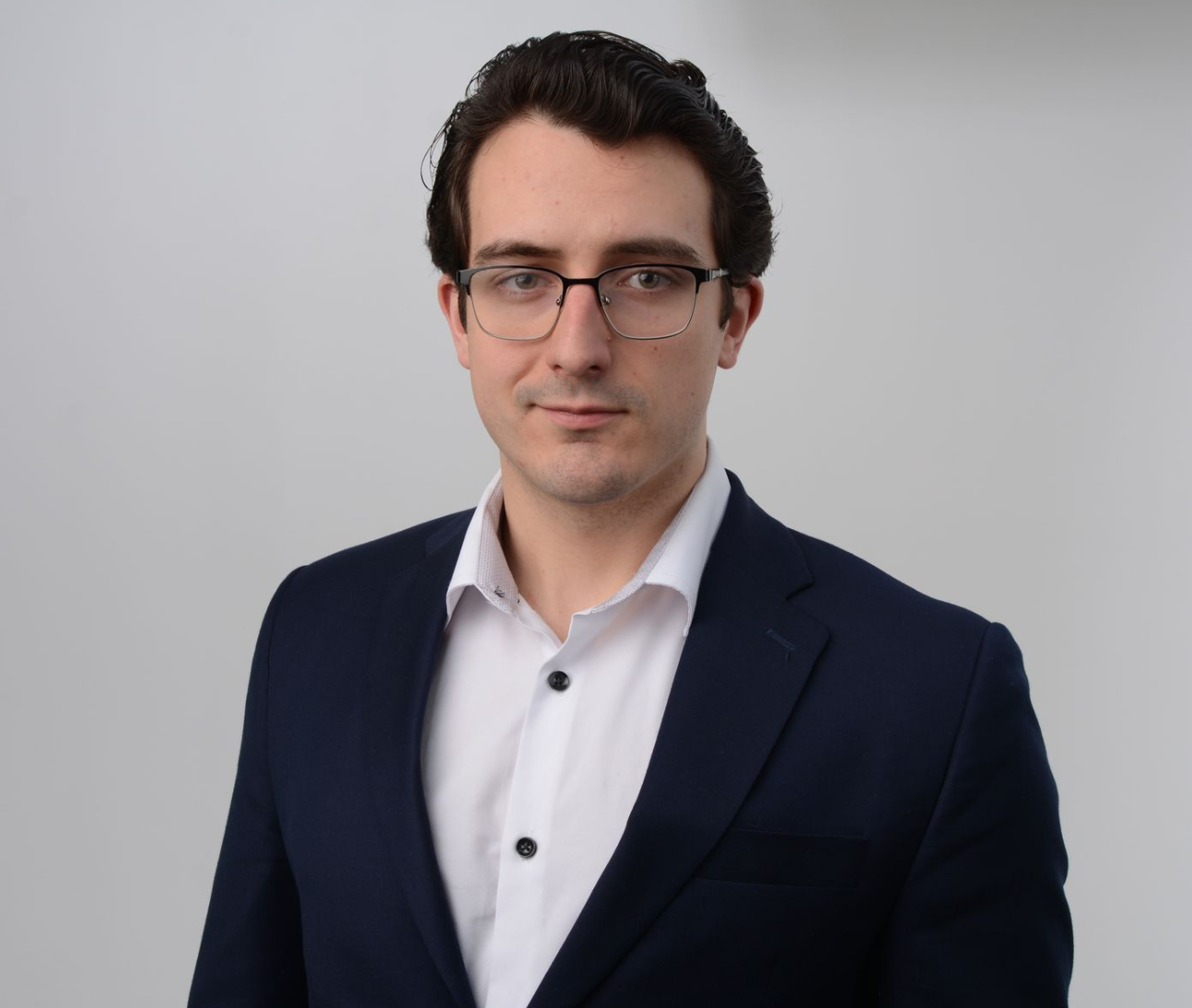 Man with glasses and a dark suit, looking at the camera. Against a white background.