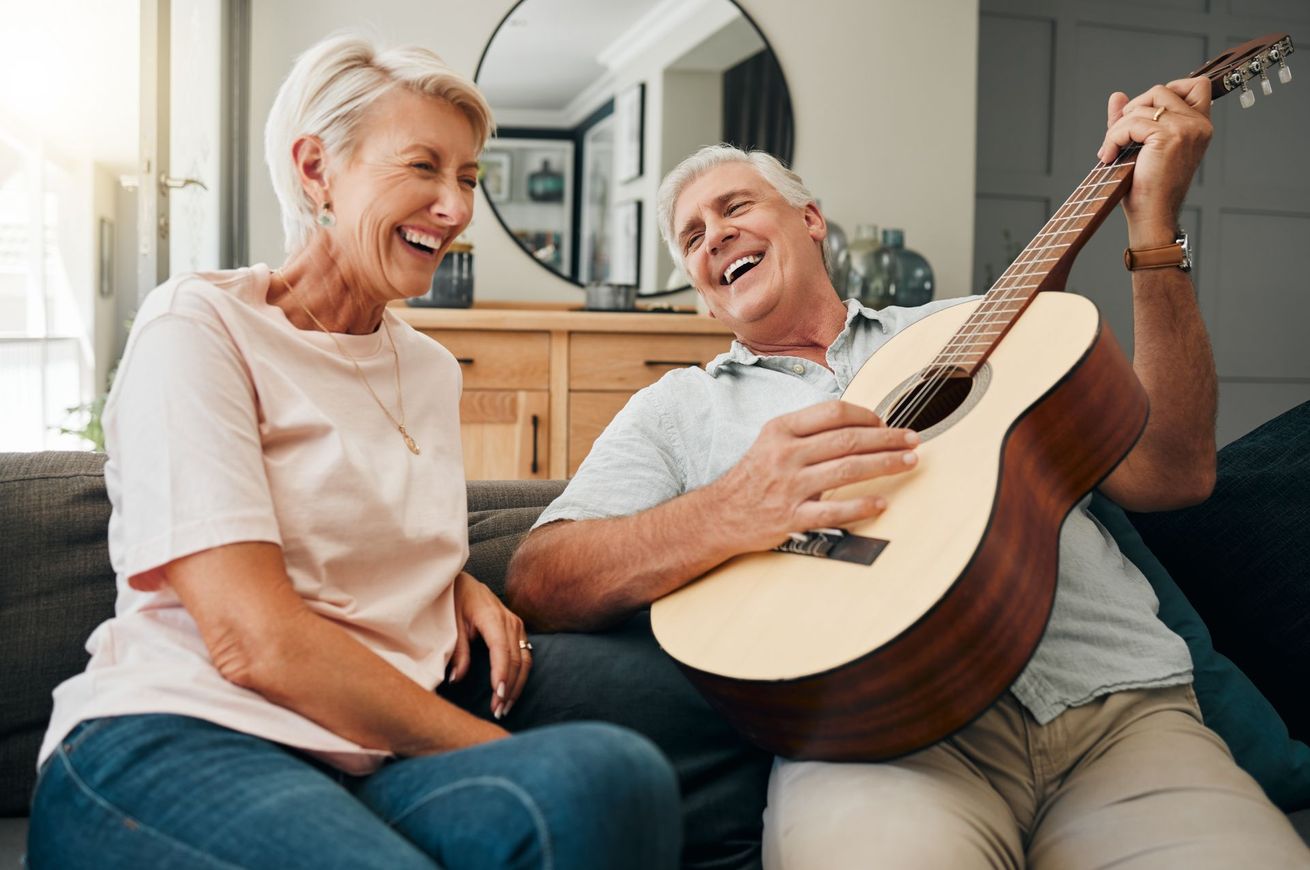 Smiling senior couple, man playing guitar, woman laughing, sitting on couch in living room.