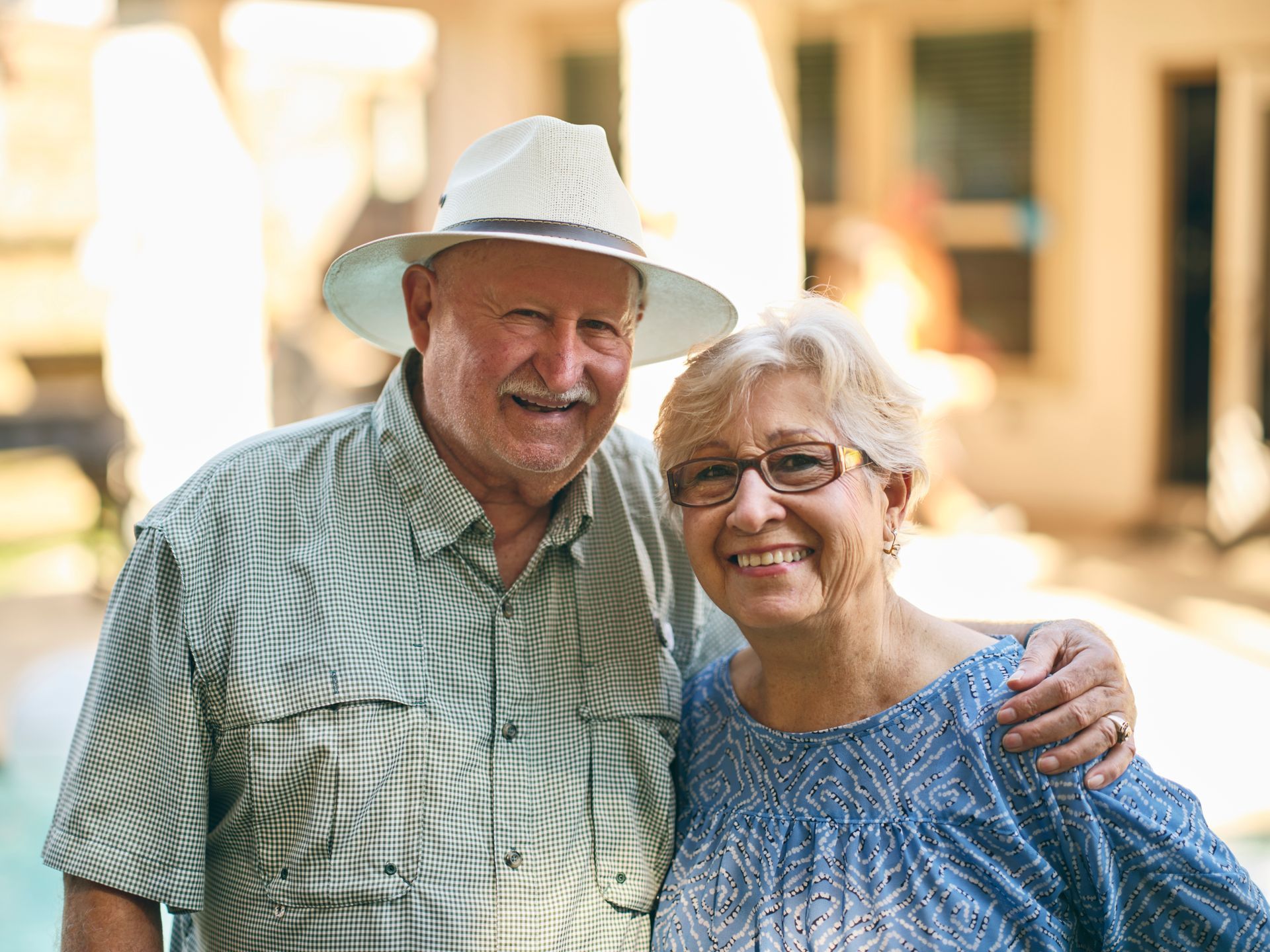 Smiling elderly couple outdoors; man with hat and arm around woman with glasses.