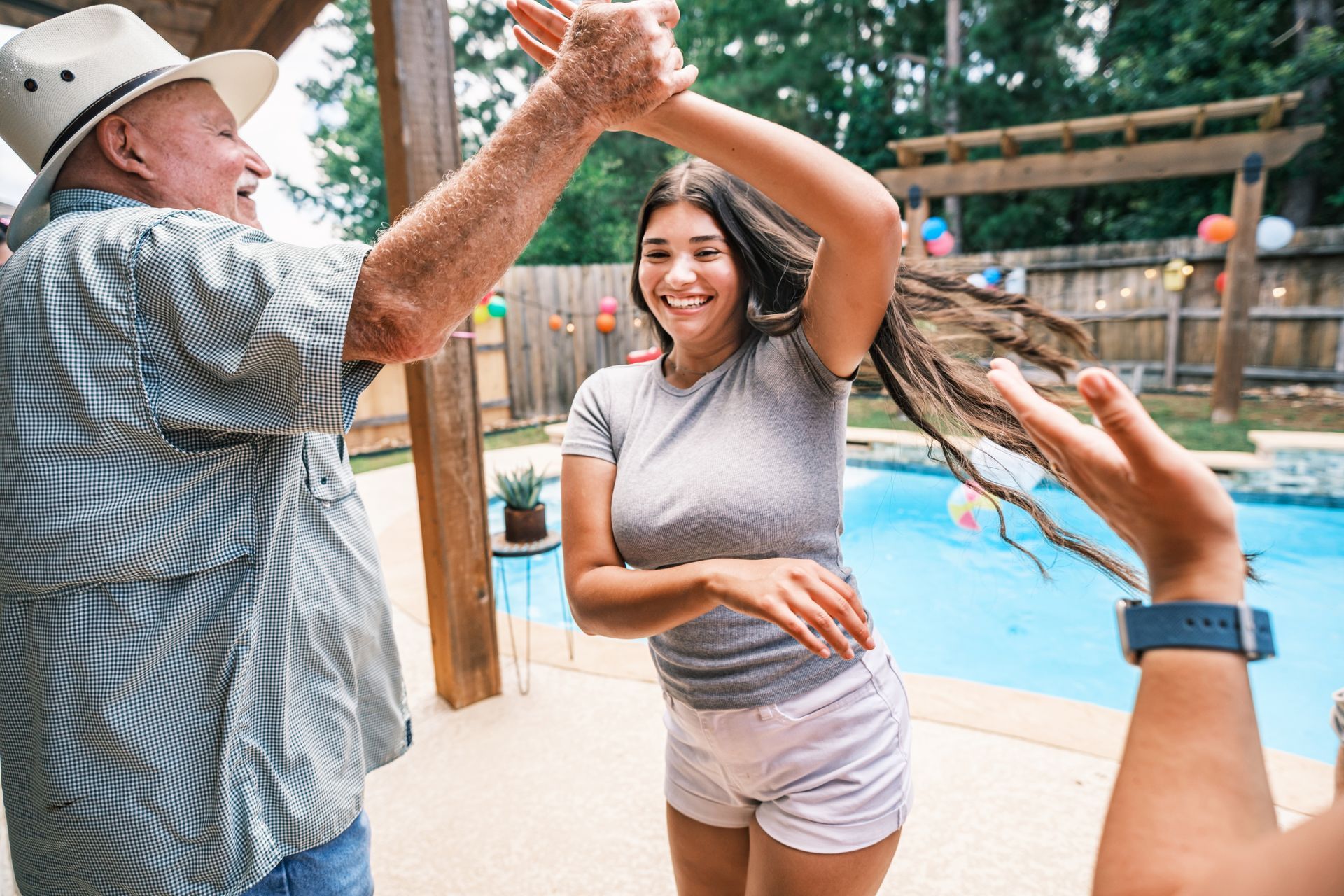 An older man and young woman dancing in a backyard, by a pool, laughing and celebrating.