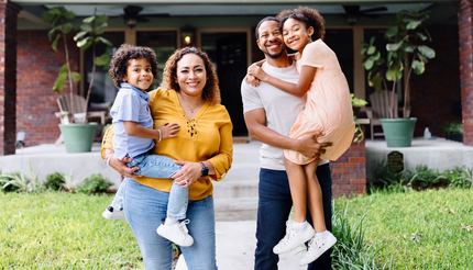 Family of four smiles in front of their brick home; mother holding son, father holding daughter.