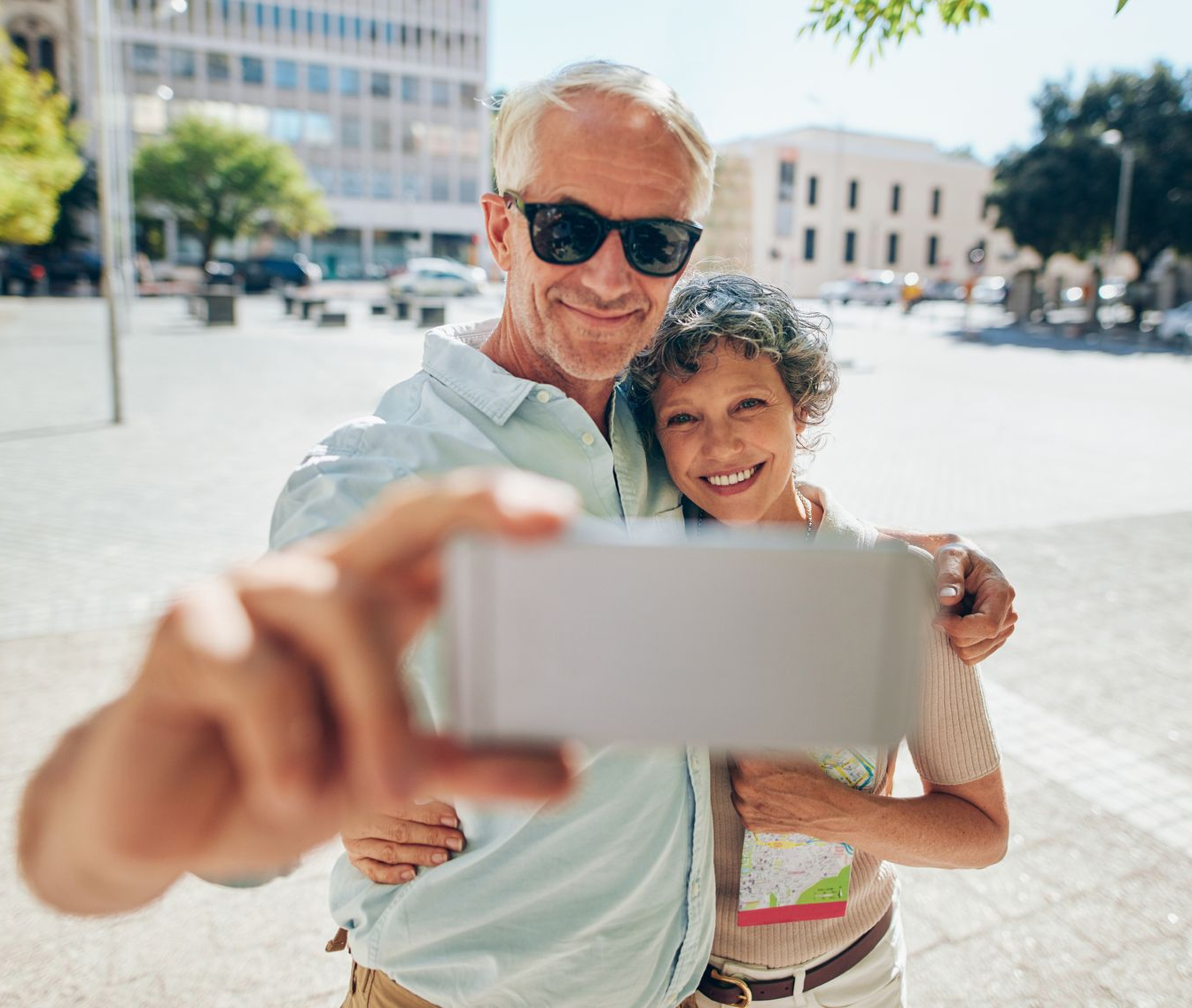 Senior couple smiling, taking a selfie outdoors. Man holds phone, woman has a map. Sunny day in a city square.