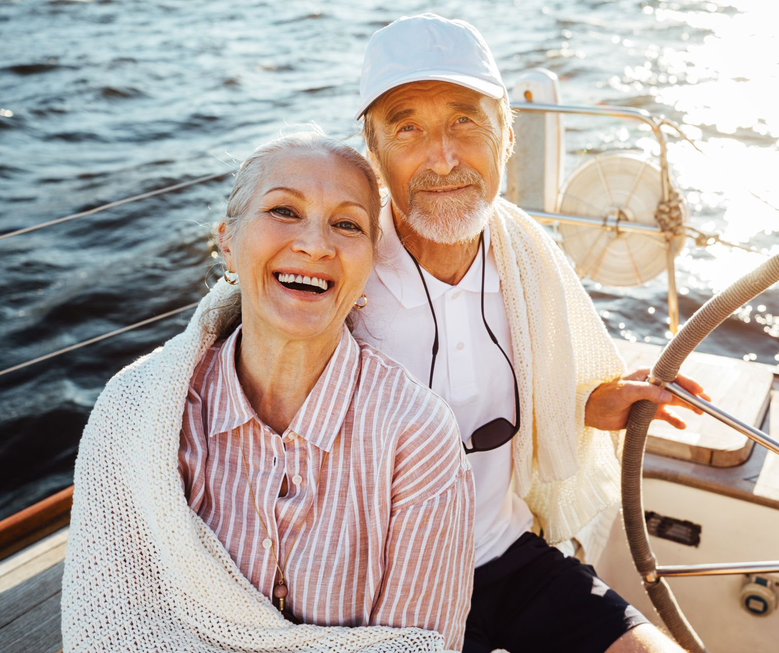 Smiling elderly couple on a boat, woman wrapped in blanket, man at the wheel, water and sun in the background.