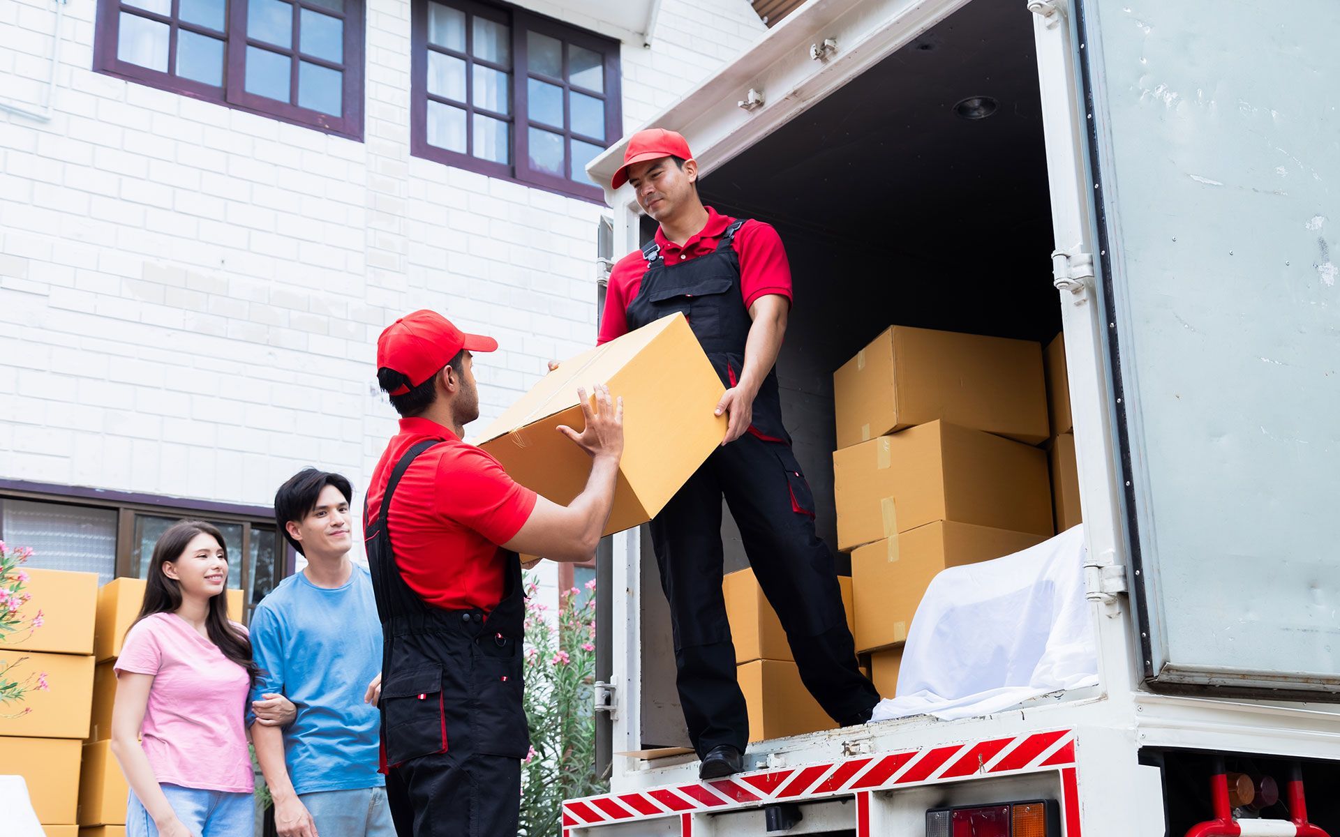 Two movers unloading boxes from a truck for a couple; red uniforms, outdoor setting.