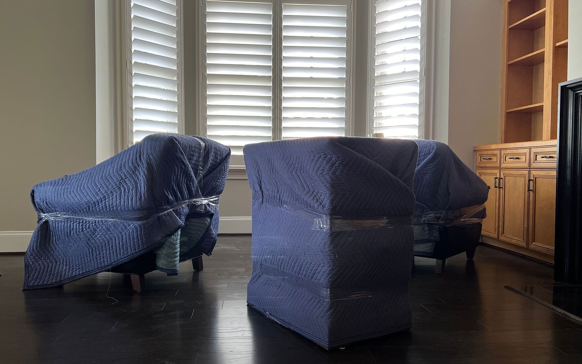 Two blue-covered chairs and one table are in front of a window with closed blinds. Cabinets are in the background.