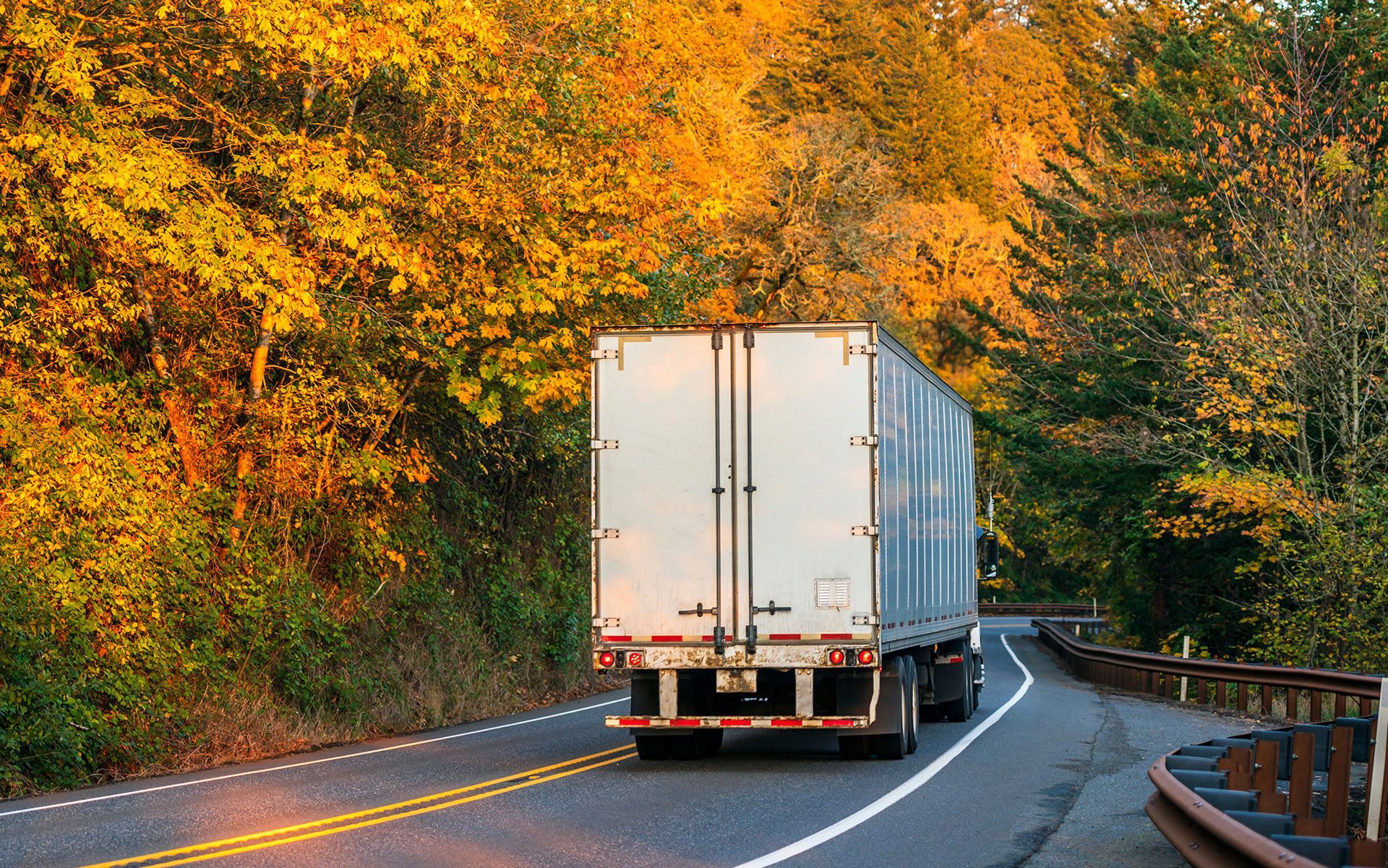 Semi-truck on a curved road, autumn trees with yellow and orange leaves in the background.