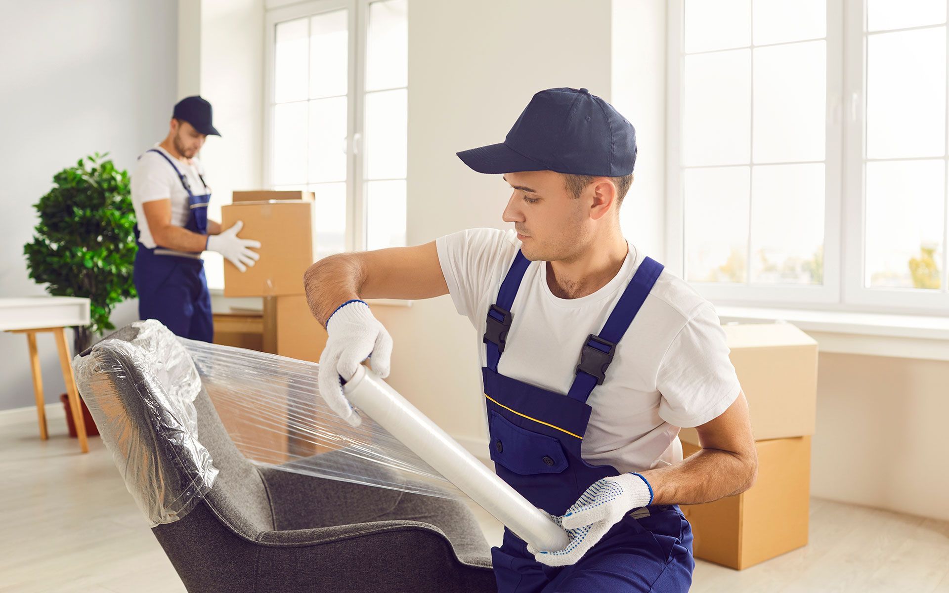 Movers wrapping chair with plastic wrap in a brightly lit room with boxes. Another mover in background.