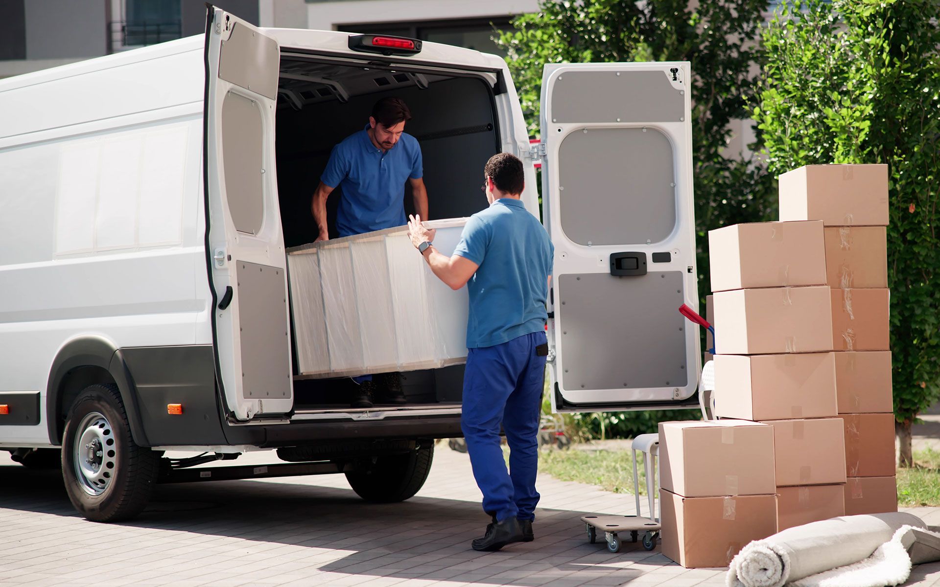 Two movers loading a mattress into a white cargo van next to a stack of boxes on a sunny day.