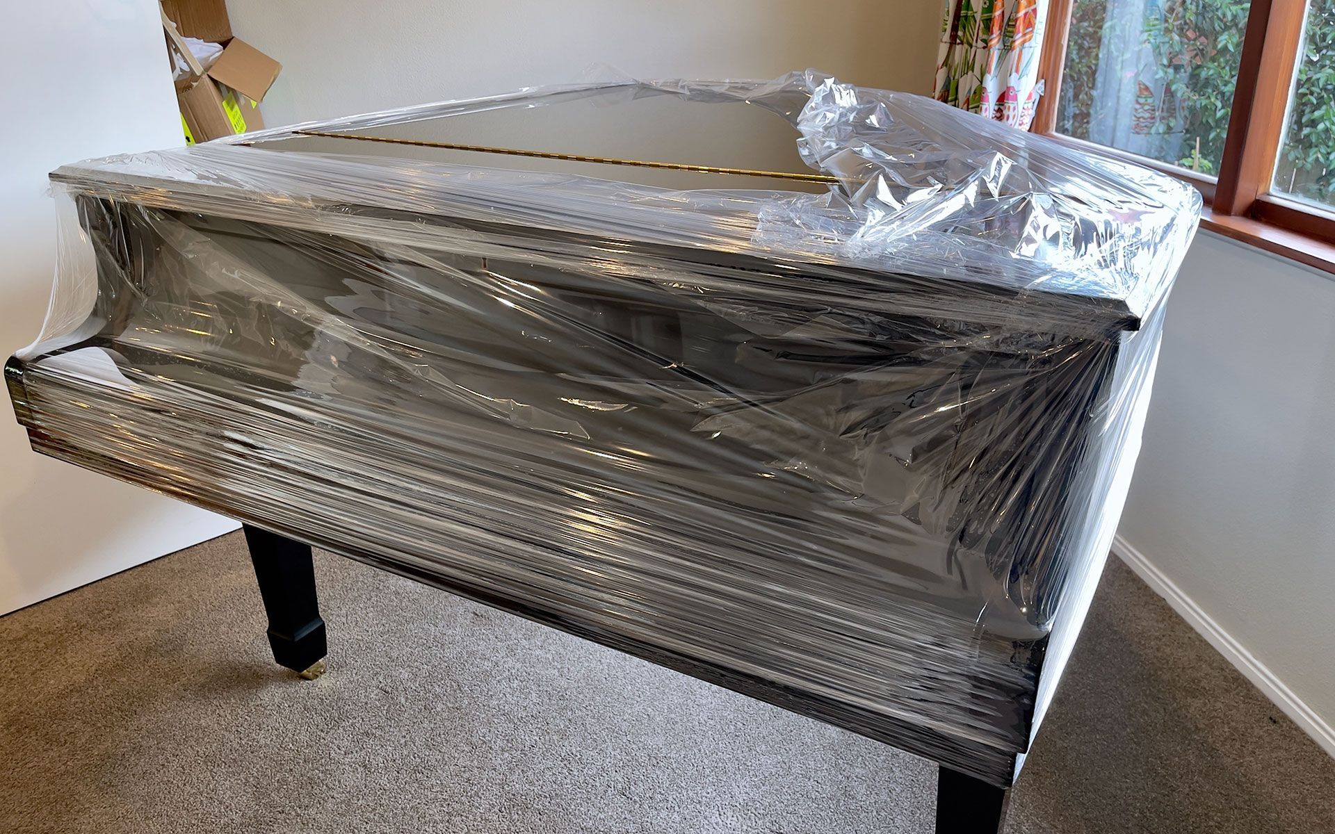 Piano wrapped in plastic, ready for moving, near a window and cardboard box.