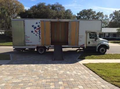 Moving truck parked on a paved driveway with ramp extended; doors open, street and houses in background.