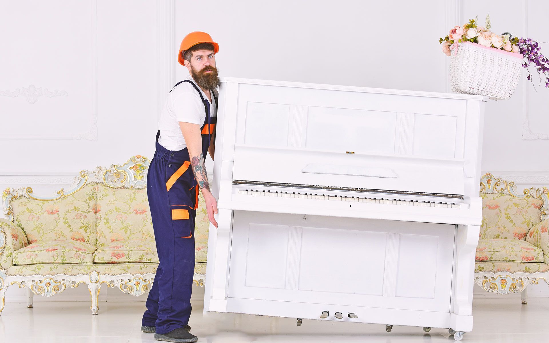 Bearded person in overalls and hardhat lifting a white piano in a room with a floral couch.