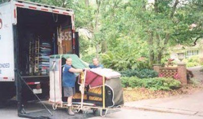 Two movers loading a piano onto a truck. Green and brown tones. Trees and house in the background.