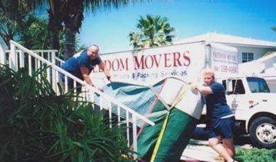 Two movers loading a wrapped couch down outdoor stairs toward a moving truck.