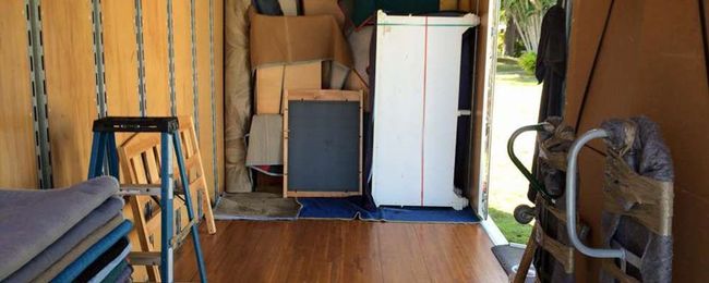 Interior view of a cluttered trailer. Contains boxes, furniture, a ladder, and a chalkboard. Wooden floor and tan walls.