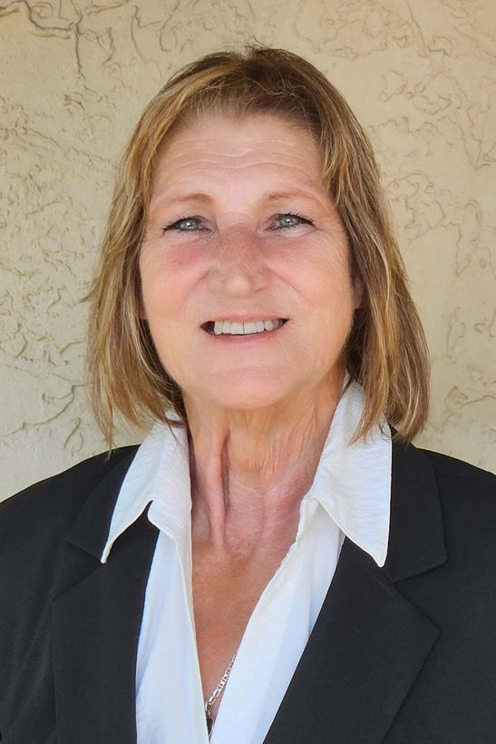 Woman with blond hair, wearing a white shirt and black blazer, smiling in front of a neutral wall.