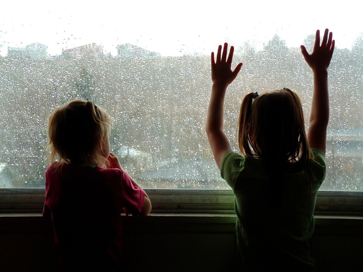 Two children looking out a window at the rain. One child has hands up.