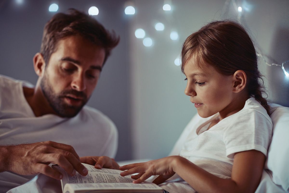 Man and child in bed reading a book together under fairy lights.