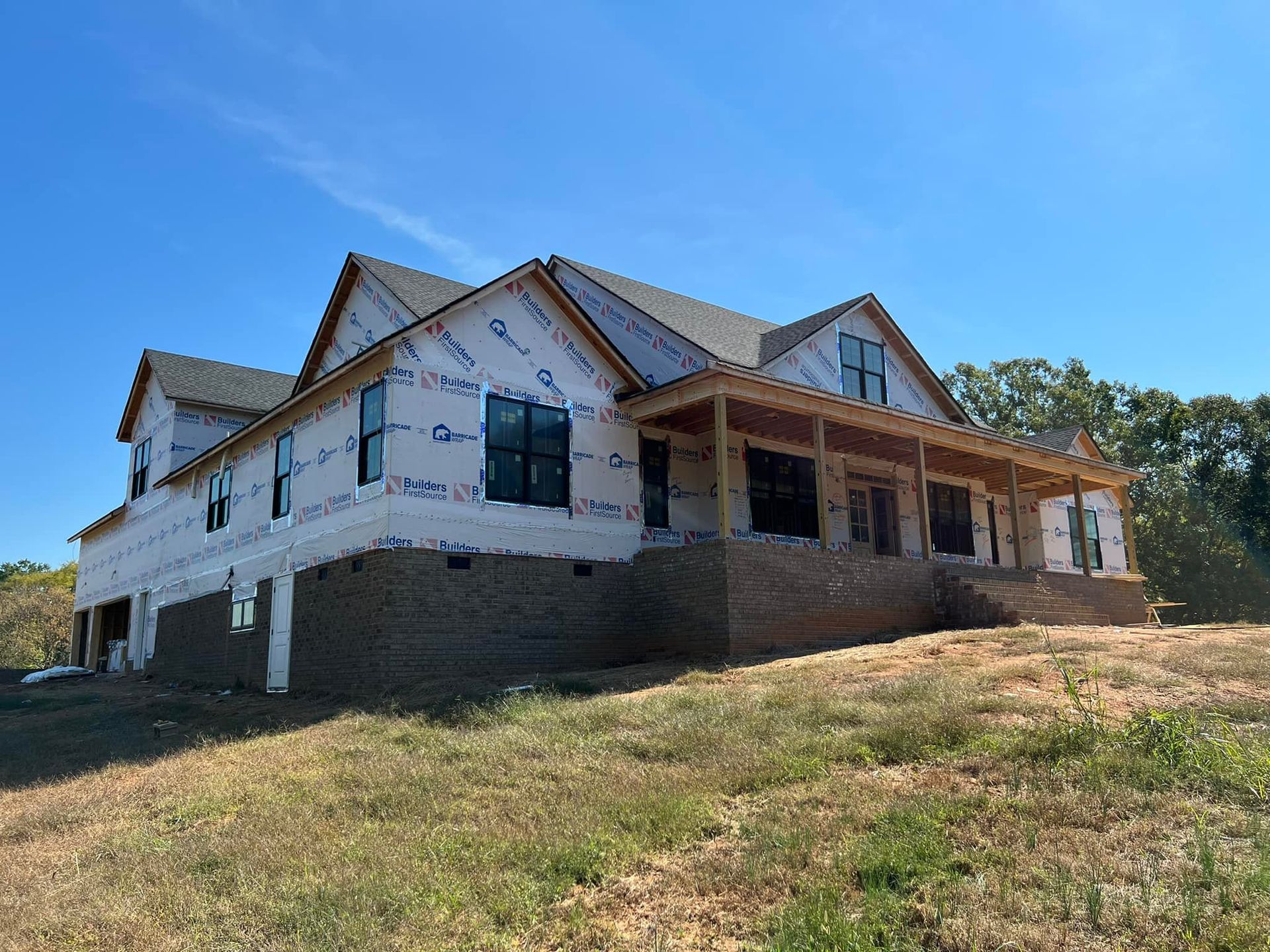 New house under construction on a grassy hill; blue wrap, dark windows, and a porch.