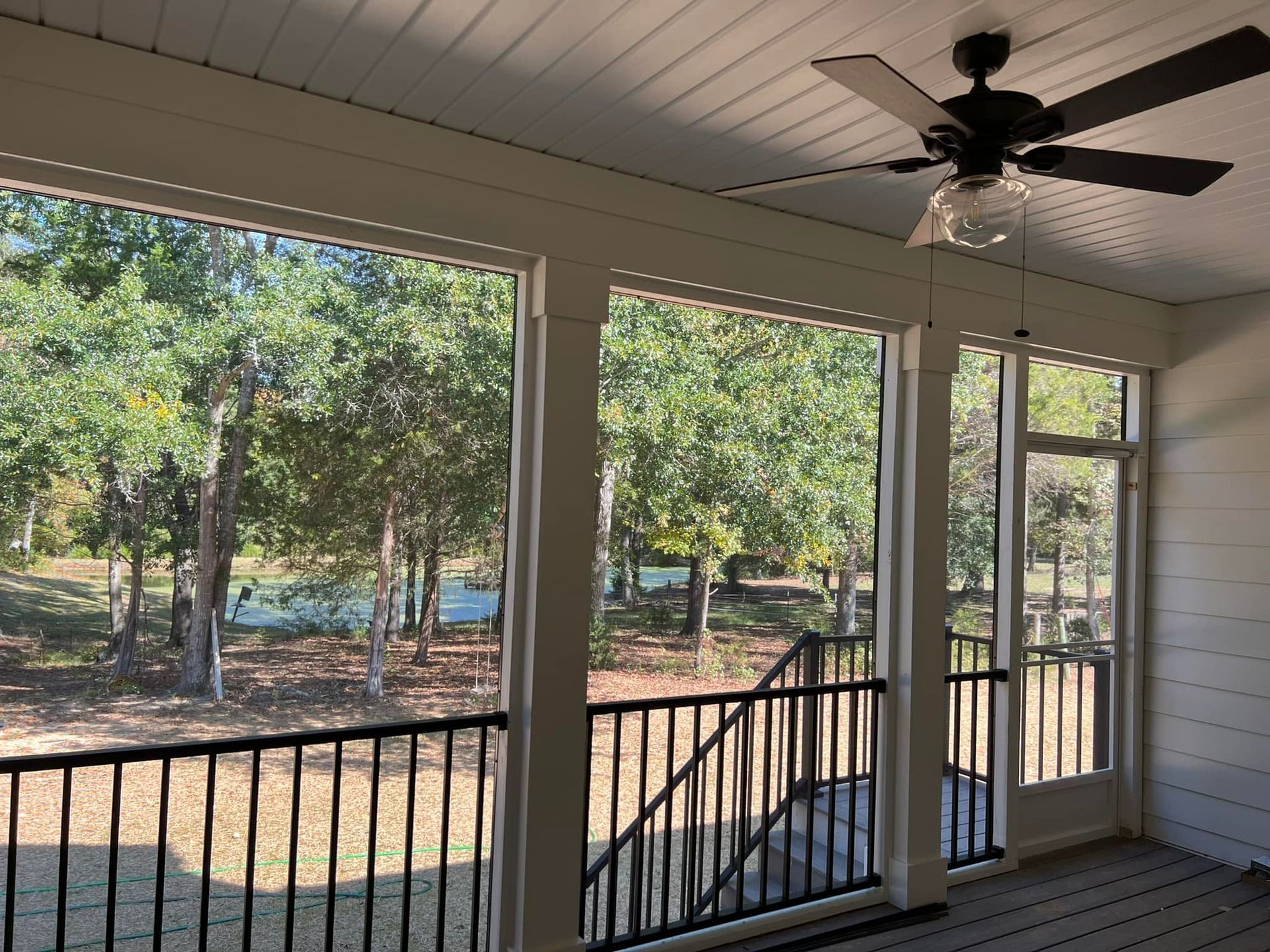 Screened porch with black railing, white trim, and ceiling fan overlooking a wooded area.