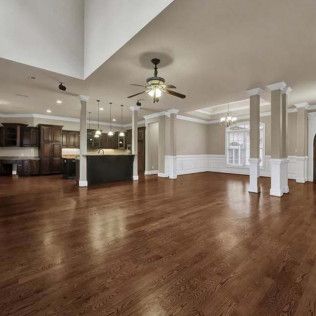 Spacious living area with dark wood floors, kitchen in the background, columns, and a ceiling fan.