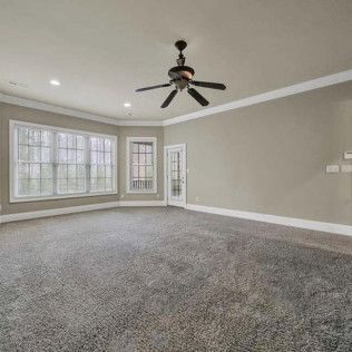Empty room with gray carpet, walls, and trim. Ceiling fan hangs from the ceiling. Windows and door.