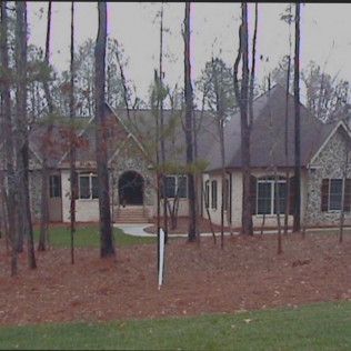 A beige, stone-and-wood house sits amidst trees on a lawn. Gray sky overhead.