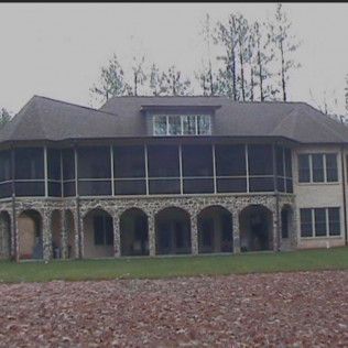 Large house with stone arches, screened porch, and brown roof. Green lawn in front.
