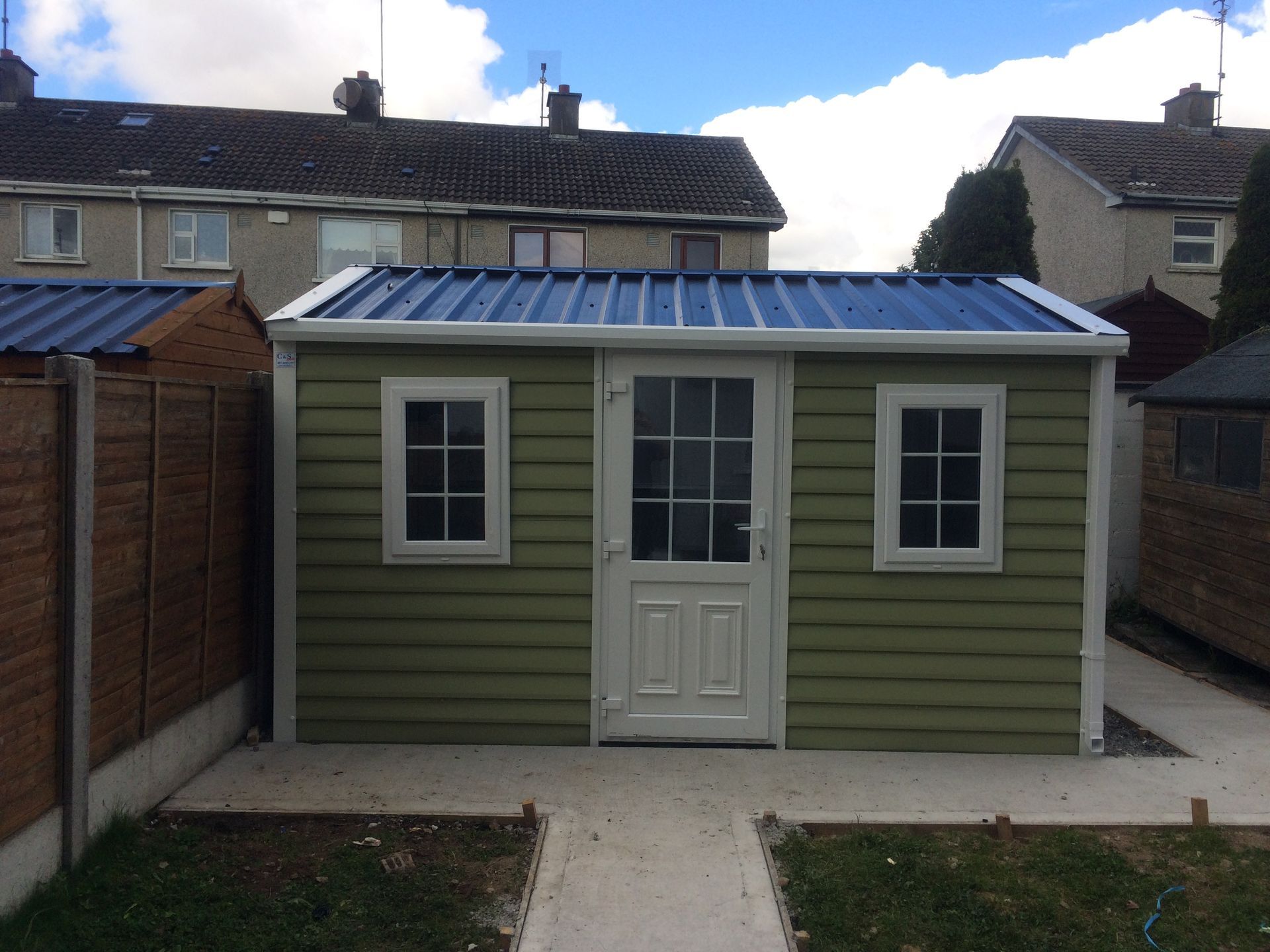 Green shed with white trim, blue corrugated roof, and a white door with two windows.