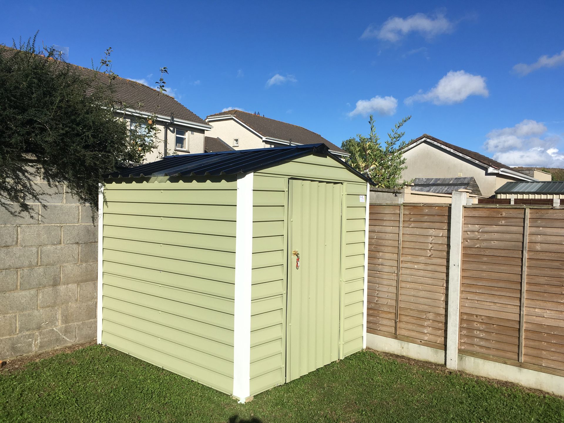 Green shed in a backyard with a wooden fence and houses in the background under a blue sky.