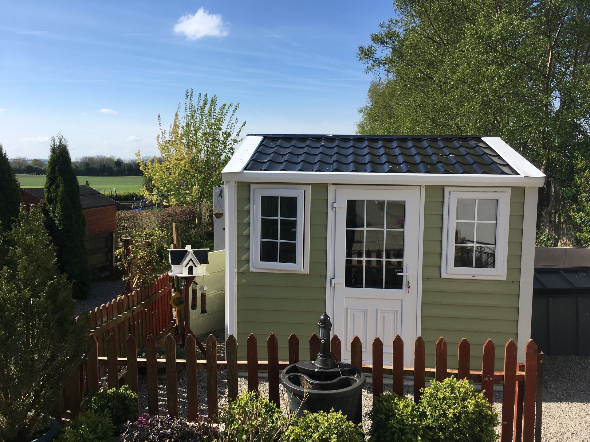 Green garden shed with white trim, black roof, and picket fence, set against a sunny landscape.