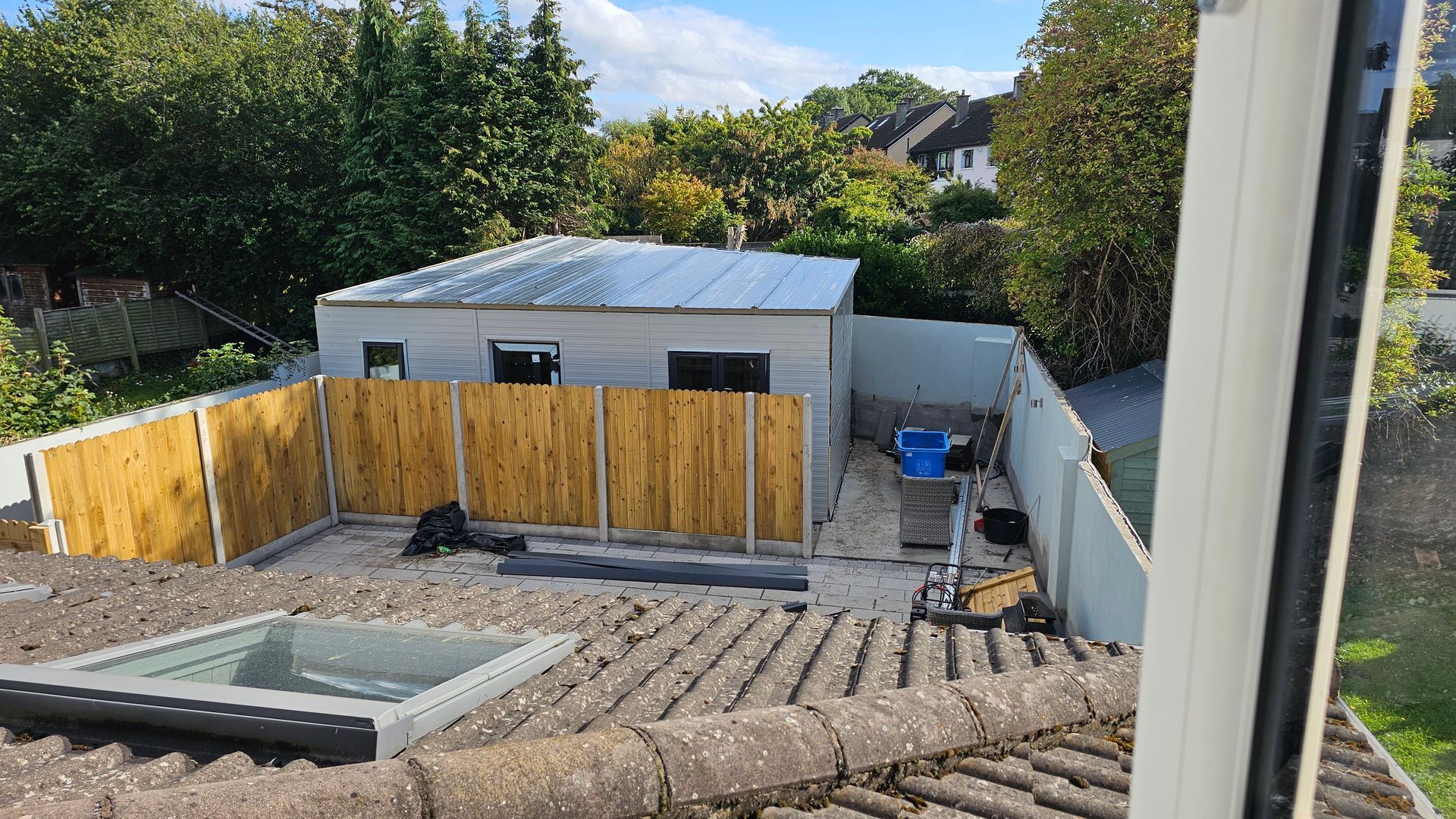 Backyard with shed, wooden fence, and tiled roof. Blue sky, green trees, and a skylight.