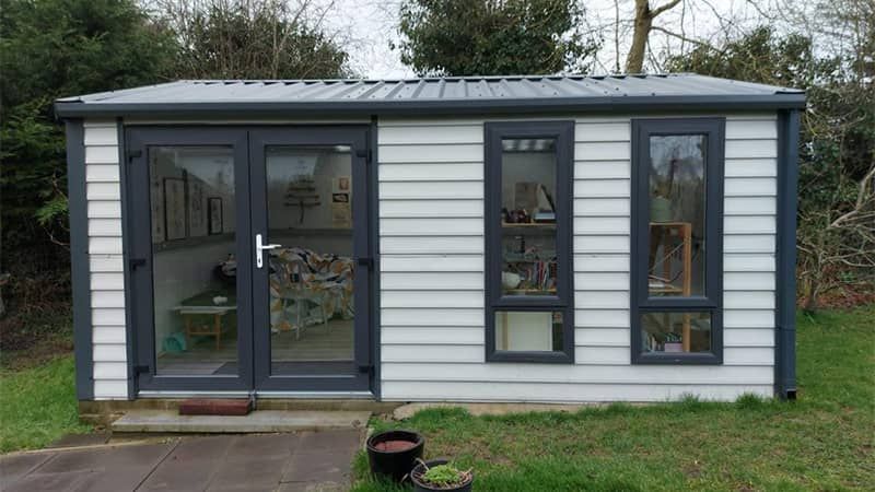 A small, white garden shed with gray trim, windows, and double doors; surrounded by greenery.