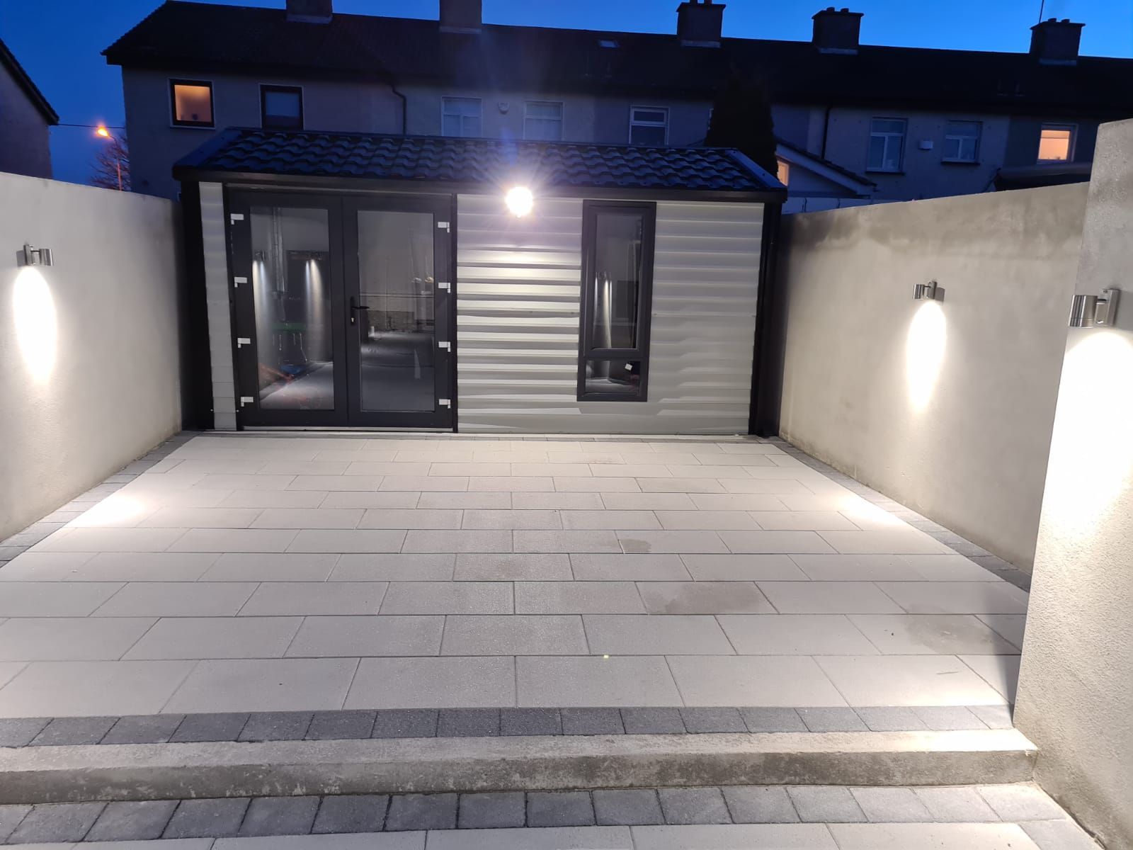 Lit outdoor patio with building, featuring concrete pavers, white walls, and spotlights at night.