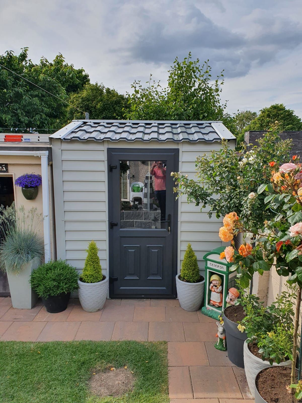 Gray shed with dark gray door, surrounded by potted plants and a red brick patio.