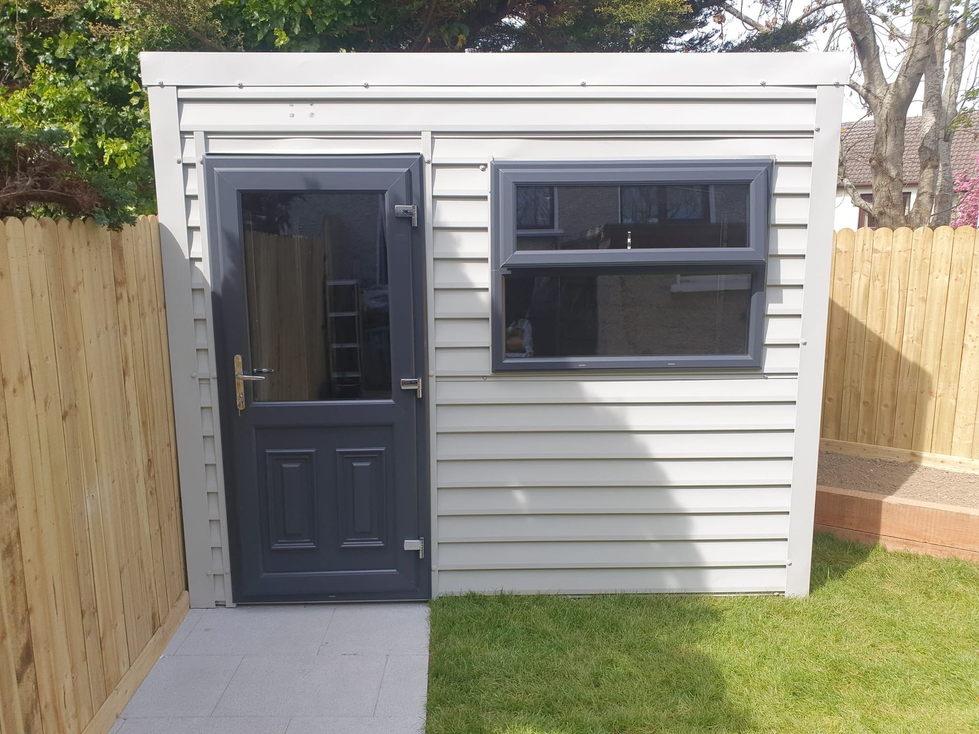 Grey shed with dark grey door and window, on a grassy lawn beside a wooden fence.