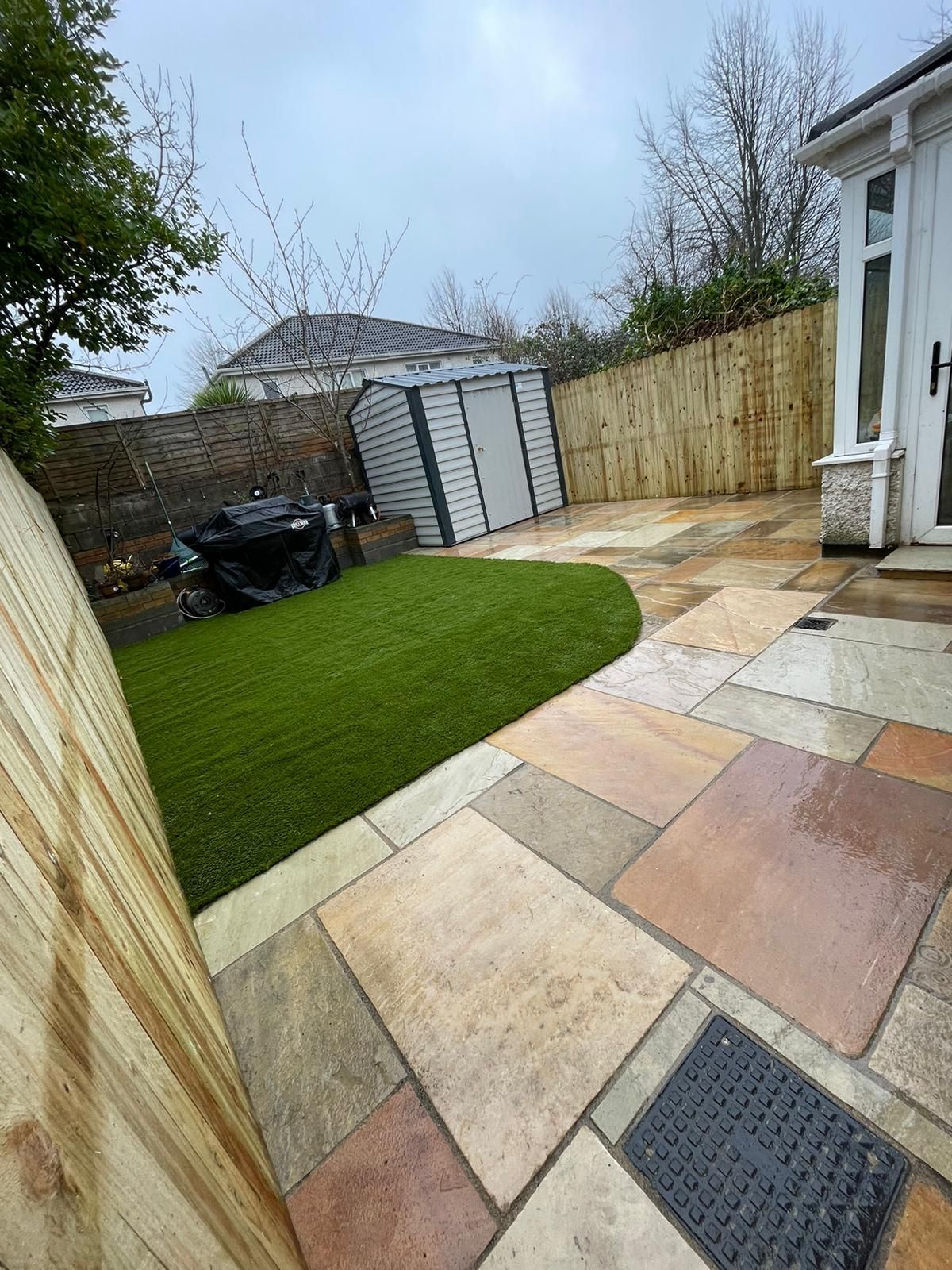 Backyard with a patio, artificial lawn, shed, and wooden fence. Overcast day.