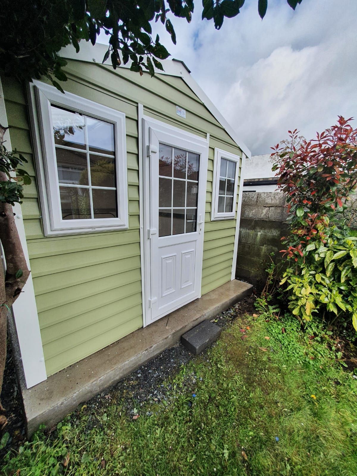 Green shed with white door and window frames; set in a grassy area with greenery and cloudy sky.