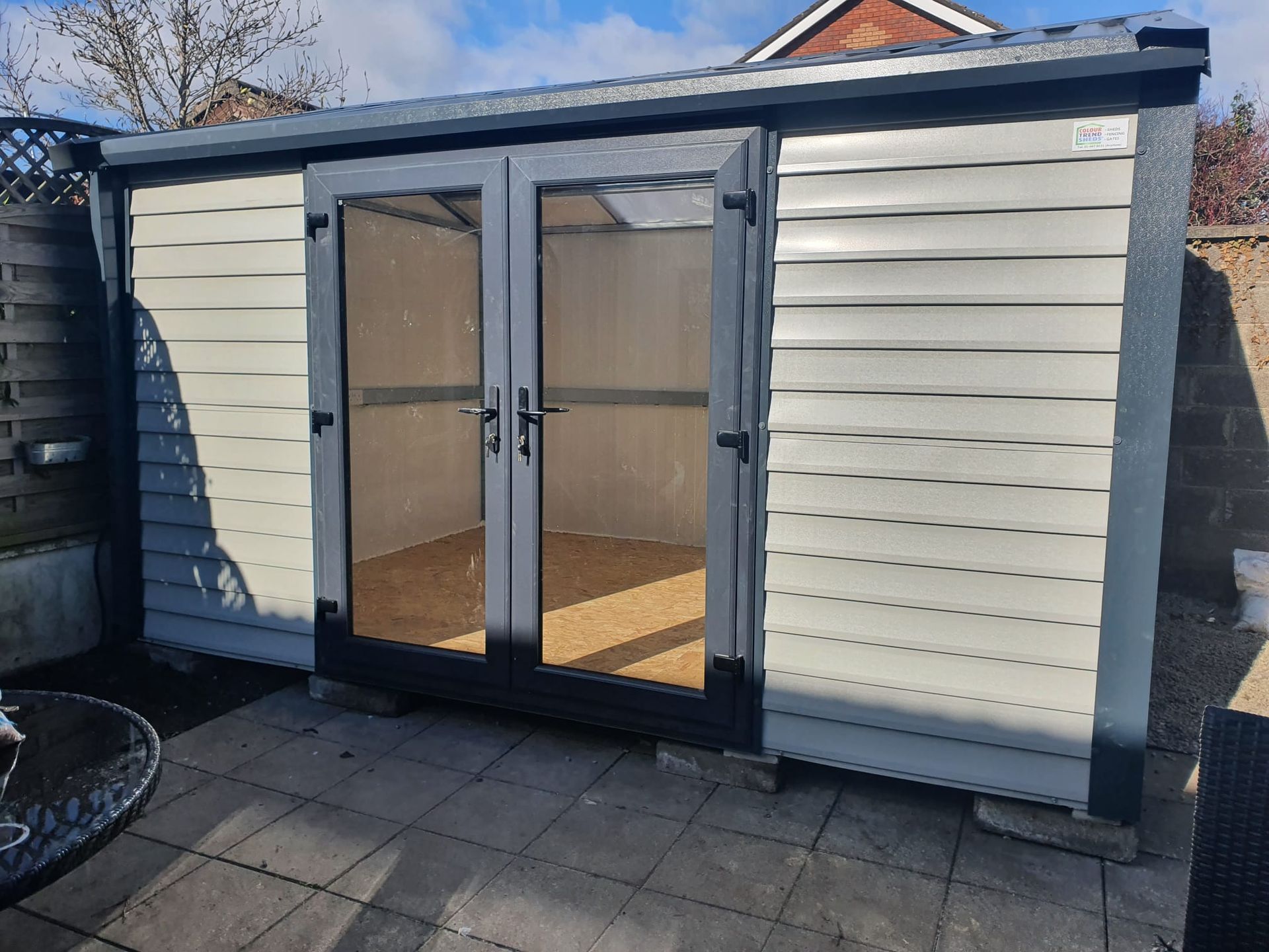 Gray and cream shed with double glass doors on a paved patio, against a brick wall.