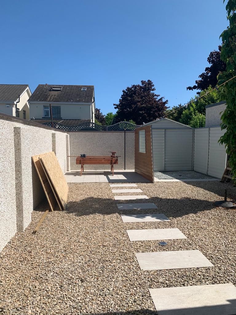 Gravel yard with stepping stones, shed, and a wooden bench. The yard is enclosed by stucco walls, with a clear blue sky.