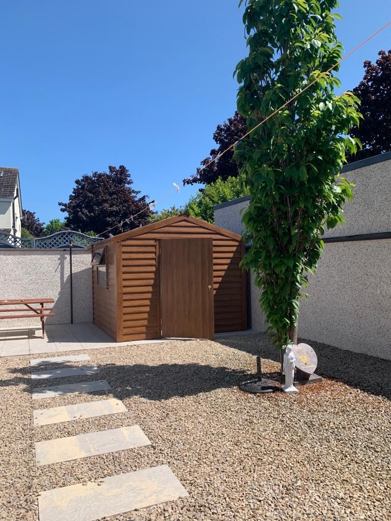 Brown shed in a yard with a stone path, gravel, and a tall tree under a blue sky.