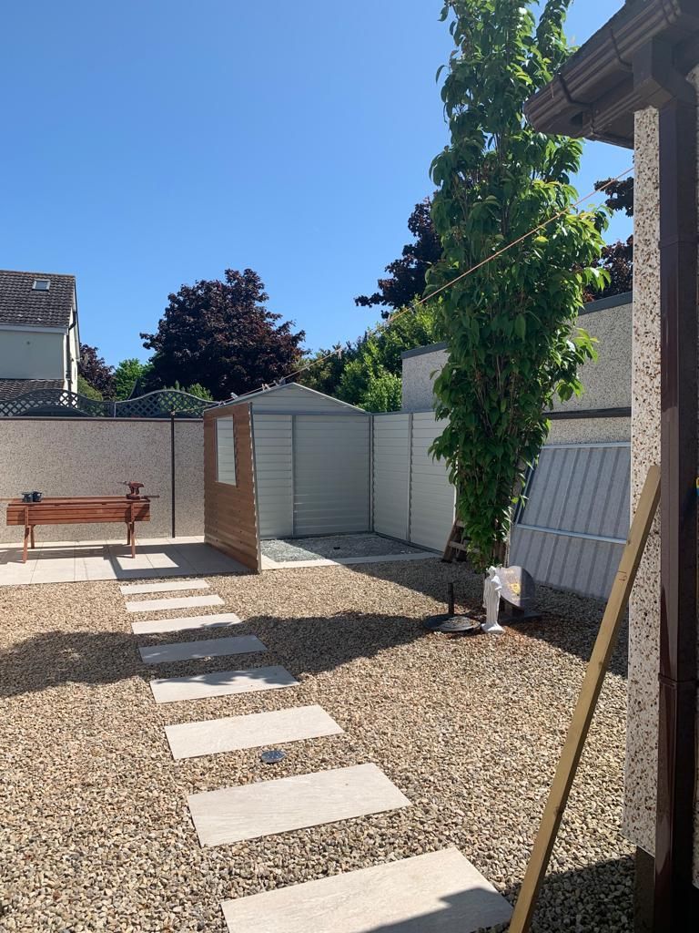 Stone pathway in a gravel yard leading to a shed. A tree, walls, and structures frame the space under a blue sky.