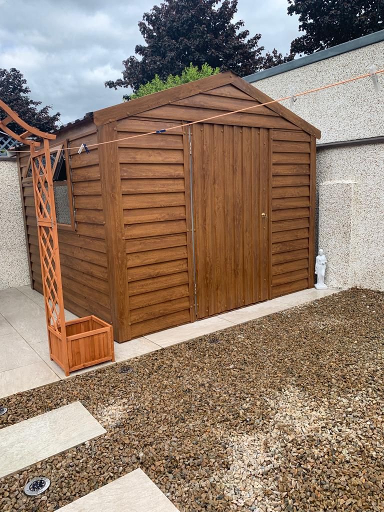 Brown wooden shed with a small trellis in a yard with gravel and pavers.