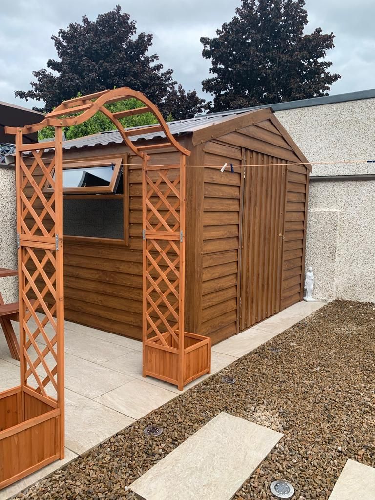 Wooden garden shed with trellis archway on patio with gravel and a dark tree background.