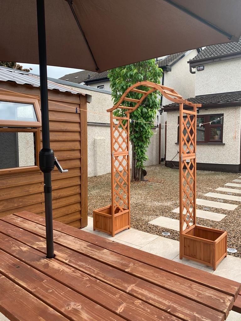 Wooden arbor with built-in planters in a backyard. Brown umbrella and wooden table in foreground.