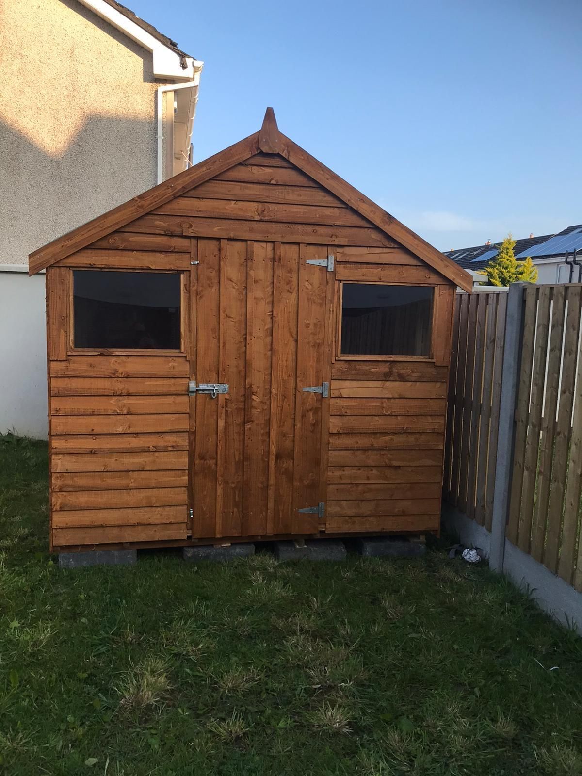 Wooden shed with two windows and double doors, set on a grassy lawn next to a fence.