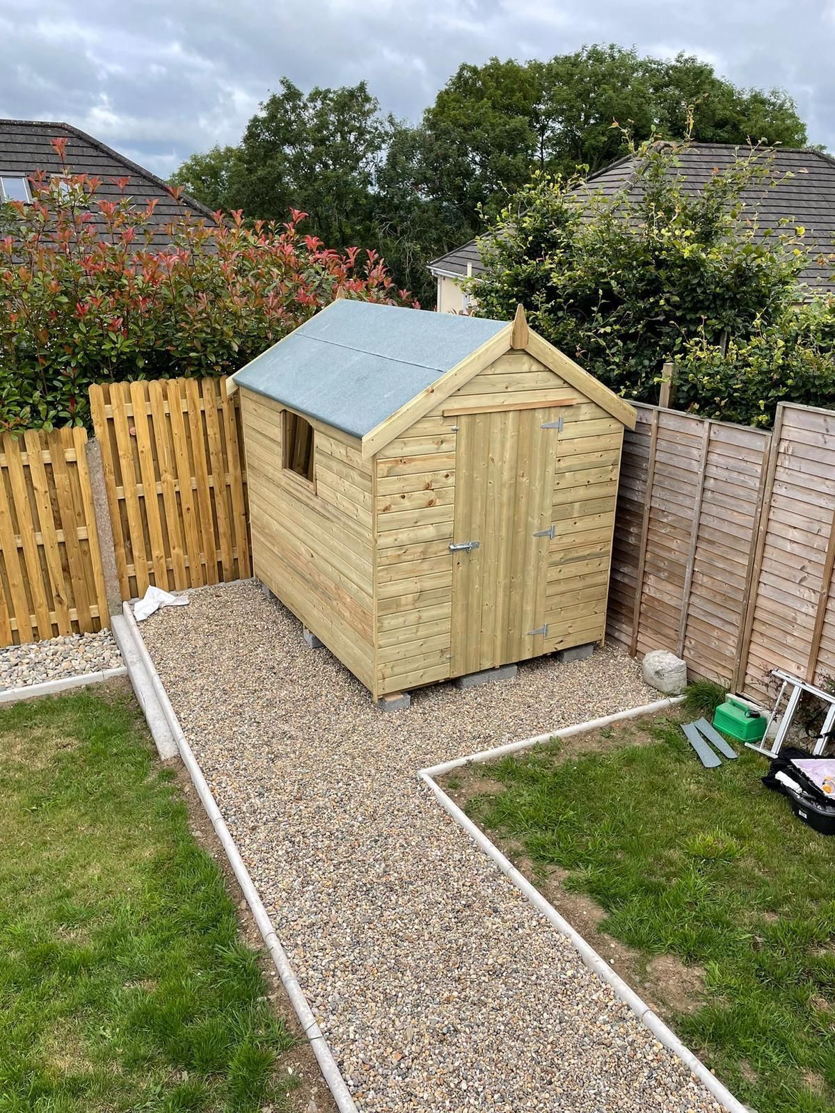 Wooden shed in a backyard with a gravel path, flanked by fences and greenery.